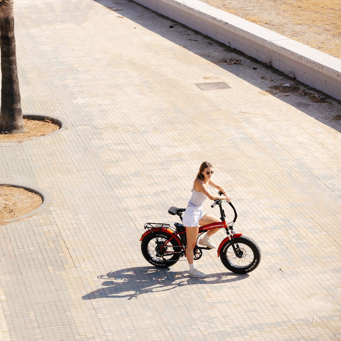 Woman riding a red folding fat tyre e-bike across a sunny paved square