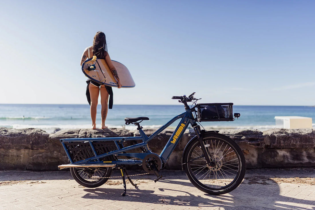 Woman carrying a surfboard beside a blue longtail cargo e-bike by the sea